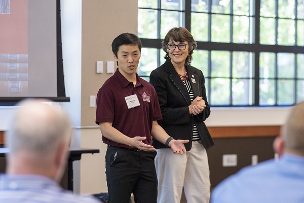 Chico State President Gayle Hutchinson stands next to a male student who is making a presentation with their backs to a drop-down projector screen.