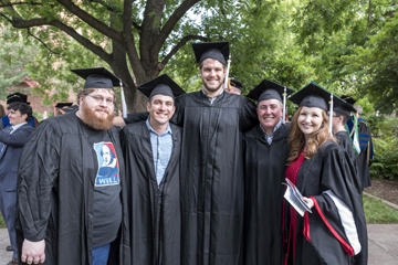 A group of five graduates (four male and one female), wearing black caps and gowns, some open and askew, pose together in a line. The women, on the right end of the line, wears a red sash and is holding a groups of papers in her left hand.