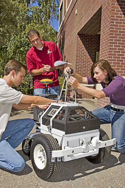 One man holding a clip board inspects a machine to which a woman and man apply finishing touches.