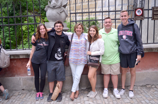 Six people stand in a line, posing against a wall with a stone statue encompassed by black bars on either side.