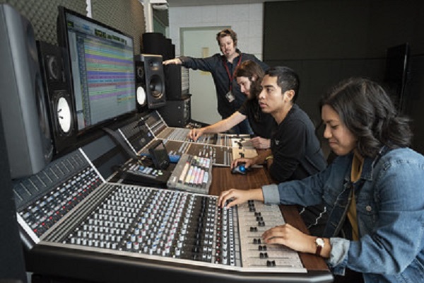 A group of three students and a teacher sit in front of a monitor as the students work the control board.  