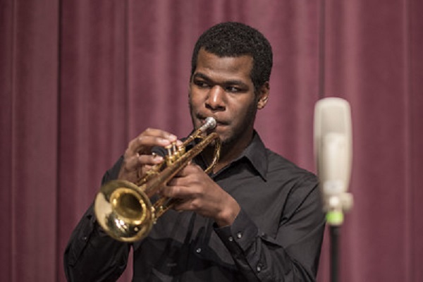 A man dressed in a black shirt plays the saxophone against a red background.