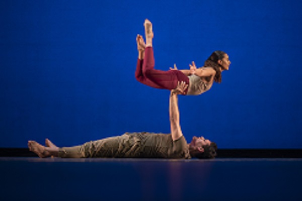 A male dancer lays flat on his back while supporting a female dancer with his hand on her hips as he lifts her above him.