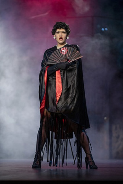 A person dressed in black stands while holding a hand fan across their chest with a black gloved hand.