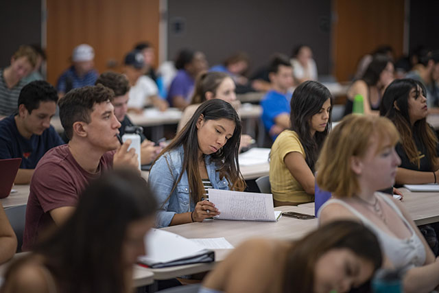 Students sit in rows of tables taking notes with the focus on one female student who has flipped up a page of her notebook and seems to be reviewing her notes.