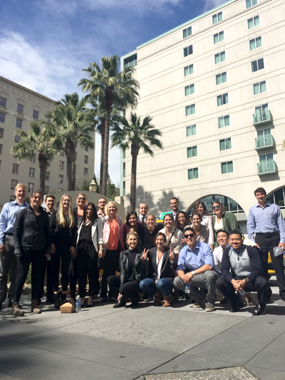 A diverse group of students, pose for a photo in front of, what appears to be, a light tan-colored hotel building--some holding up a "peace sign" with their fingers, some with water, all wear smiles.