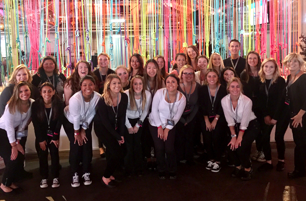 A group of people kneeling down, with hands on their knees, pose in front of a multi-colored curtain (of sorts) made from various brightly colored ribbon strands.
